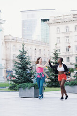 Fototapeta na wymiar Smiling friends in sportswear running in the city dicussing and giving hi five with hands. Multiethnic women having a fitness workout.