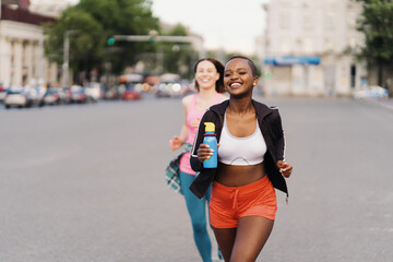 Cheerful smiling friends in sportswear running in the city dicussing. Multiethnic women having a fitness workout.