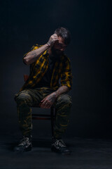 Handsome male model with tattoos and tartan shirt is sitting on a chair and is touching his hair while isolated on a black background