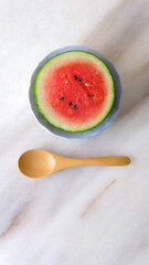 Flat lay of a small round watermelon cut in half, and a wooden spoon next to it. Placed on a marble counter top.
