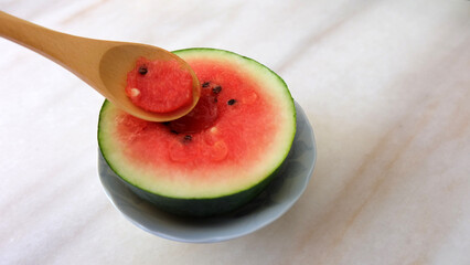 Wooden spoon scooping out some part of a small watermelon cut in half. With marble background.