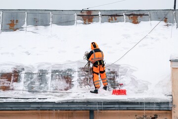 A working man in bright overalls with a safety belt with a shovel clears snow from the roof of an old building. Prevention of snow falling from the roof, industrial mountaineering - Powered by Adobe