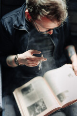 Adult student reading a book in the college library while smoking. Young male wearing glasses with bookshelves on background