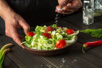 Professional chef sprinkles salt to salad of fresh vegetables on wooden table. Preparing healthy food on the kitchen