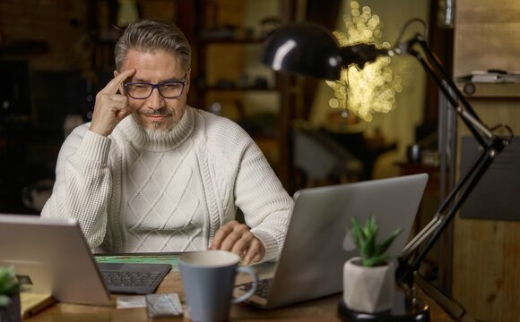 Happy mid adult man working with laptop computer in home office, glasses, gray hair. Winter, Christmas, dark cosy room.