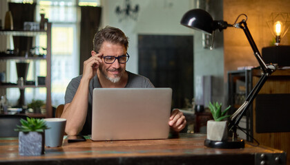 Bearded man working online with laptop computer at home sitting at desk. Home office, browsing internet. Portrait of mature age, middle age, mid adult man in 50s. - obrazy, fototapety, plakaty