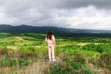 Fototapeta na wymiar Little girl in light pink clothes and long hair standing with her back on green grass in mountain valley and admiring mountains