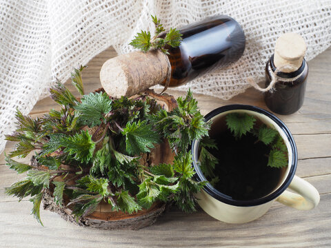 Fresh young nettle leaves, herbal drink in a mug, juice, tincture in a bottles on a wooden background, top view. Medicinal plants urtica for use in food, medicine, homeopathy and cosmetology