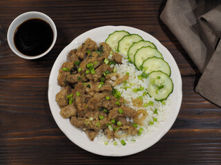 Slices of beef liver in onion sauce with garnish rice, vegetables, in a white plate, soy sauce, napkin on a dark wooden table, flat layout. Recipe for a delicious meal with byproduct for lunch