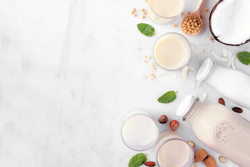 Vegan plant based non dairy milk side border. Variety of types in milk bottles and glasses with scattered ingredients. Above view over a white marble background with copy space.