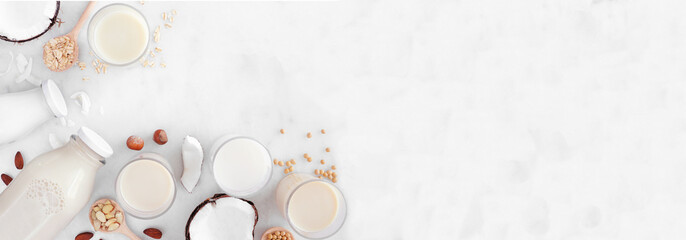 Vegan plant based non dairy milk corner border. Various types in milk bottles and glasses with scattered ingredients. Top down view over a white marble background with copy space.