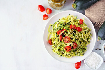 Zucchini pasta topped with tomatoes and basil pesto. Overhead view table scene on a white marble...