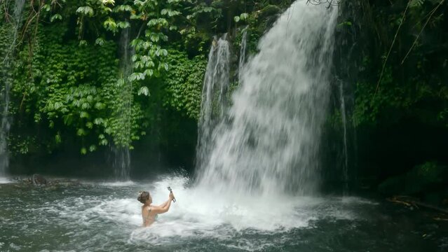 A crazy female blogger takes video on her phone under a large waterfall in tropical river. Girl with white skin under strong stream of water using modern mobile phone camera. Woman having fun in Bali