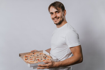 A man in a white t-shirt on a white background holds a pizza in a box.