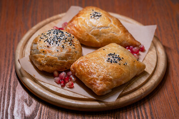 Samsa savoury pastry of Central Asian cuisines baked or fried represents a bun stuffed with meat served on round board with pomegranate seeds on dark brown wooden table at restaurant as snack