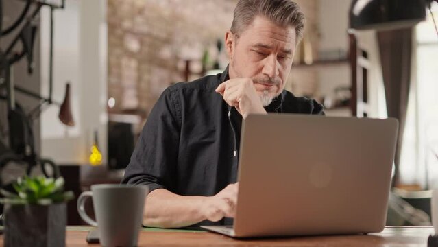 Older man sitting at desk in dark cosy room working on laptop computer in home office. Mature age, middle age, mid adult casual man in 50s, confident happy smiling.