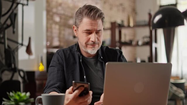 Bearded man working online with laptop computer at home sitting at desk. Home office, browsing internet. Portrait of mature age, middle age, mid adult man in 50s.