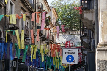 Fasce di stoffe colorate appese a dei fili tesi tra i palazzi caratteristici del centro storico di Catania, con un segnale stradale con la scritta AREA PEDONALE