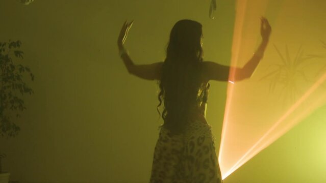 Silhouette of a woman in an exotic costume performing a belly dance, moving her half-naked body. Shot in a dark studio with smoke and yellow neon lighting.
