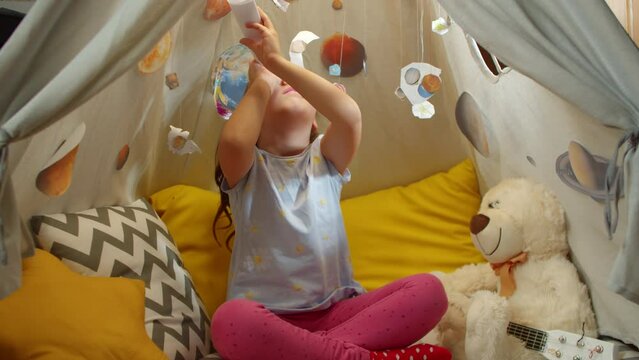 Little girl plays the explorer of space and planets sitting in a tent in the home interior. Child watches the planets through a paper telescope while sitting in a tent at home.