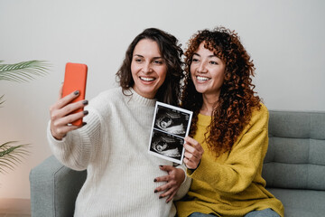 LGBT lesbian couple holding ultrasound photo scan on video call of growing baby in pregnancy time -...