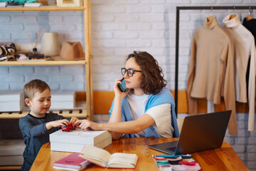 Young mother works with the little boy. Business Woman talking on the phone and discussing work issues while looking after her child. Work during coronavirus outbreak. Work remotely.