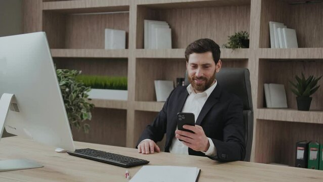 Successful handsome executive manager in formal suit looking at smartphone and smiling while reading messages or good news, sitting at his work office desk