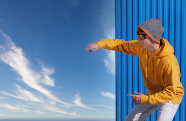 CHEERFUL YOUNG MAN IN YELLOW SWEATSHIRT, JEANS AND WOOL HAT POINTING TO THE BLUE SKY WITH CLOUDS