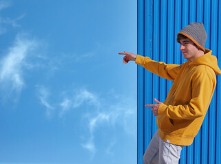 CHEERFUL YOUNG MAN IN YELLOW SWEATSHIRT, JEANS AND WOOL HAT POINTING TO THE BLUE SKY WITH CLOUDS