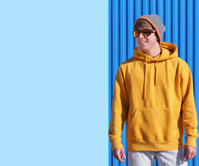 CHEERFUL YOUNG MAN IN YELLOW SWEATSHIRT, JEANS AND WOOL HAT POINTING TO THE BLUE SKY WITH CLOUDS