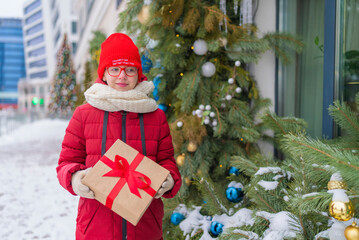 girl with a gift in red jacket and hat standing near a large Christmas tree.