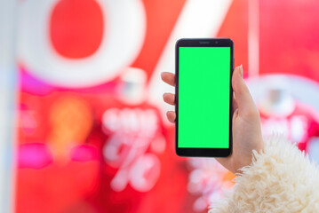 woman's hand holds a phone with a green screen to replace the background in the mall with the background of a shop window with discounts during a sale