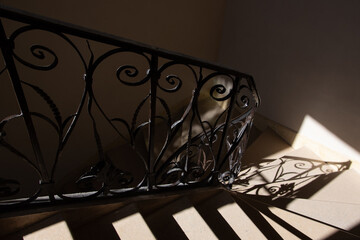 Ornate antique forging railing in sunlight rays coming from window at its silhouette reflected on stone stairs and wall. Game of light and shadow. Secret place concept. Architecture element background