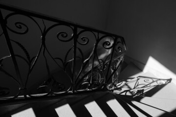 Ornate antique forging railing in sunlight rays coming from window at its silhouette reflected on stone stairs and wall. Game of light and shadow. Secret place concept. Black white historic photo