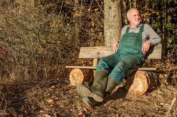 A farmerwith green dungarees and rubber boots relaxes after work on his bench at the edge of the...