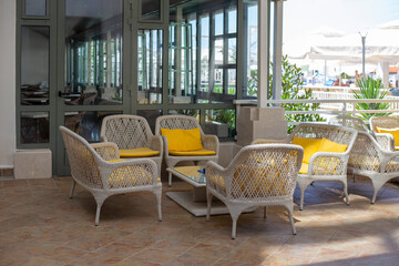 Wicker chairs with yellow cushions and tables on the terrace at summer day. Empty outdoor cafe