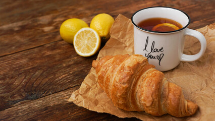 Fresh croissants and a cup of tea with lemon on a wooden table. Top view. Ingredients for delicious breakfast