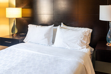 Bedroom room with illuminated lit lamp light and closeup of white pillows with sheet on mattress bed headboard in modern hotel with nobody