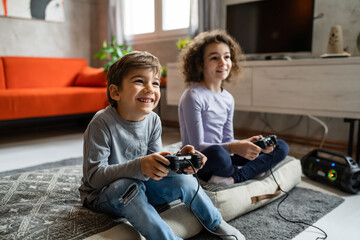 Two children small caucasian brother and sister happy children siblings boy and girl playing video game console using joystick or controller while sitting at home real people family leisure concept © Miljan Živković