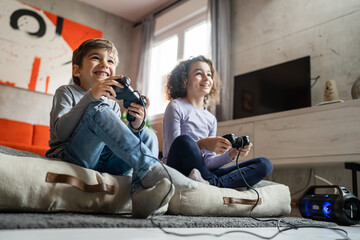 Two children small caucasian brother and sister happy children siblings boy and girl playing video game console using joystick or controller while sitting at home real people family leisure concept - Powered by Adobe