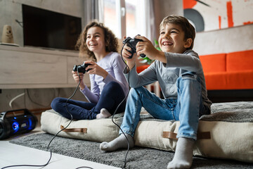 Two children small caucasian brother and sister happy children siblings boy and girl playing video game console using joystick or controller while sitting at home real people family leisure concept - obrazy, fototapety, plakaty