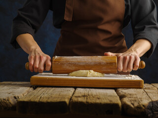 The chef rolls out the dough with a rolling pin on a wooden table, dark background. Close-up. The process of making pizza, pasta, pie. Recipes, restaurant, hotel, pizzeria, home cooking.