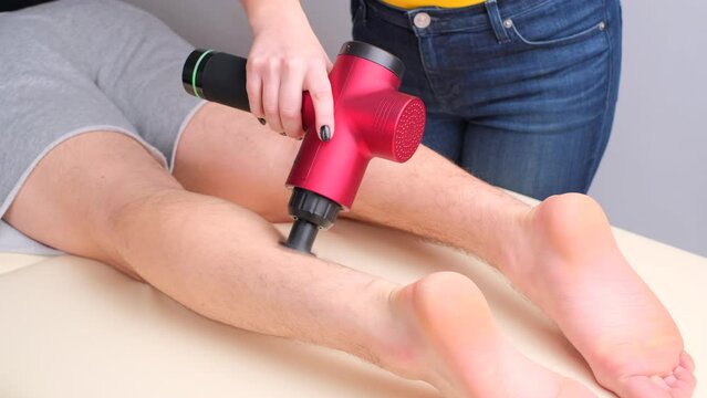 Hand of unrecognizable professional female masseur massaging calf muscles of sportsman with electric percussion vibration massager. Therapy for a regenerating massage of the athletic body 