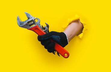 Naklejka premium The right hand, in a black knitted glove, holds several wrenches and an red adjustable wrench. Torn hole in yellow paper. The concept of a worker, a labor migrant, a master of his craft. Copy space.