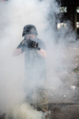 A soldier with an ak-47 in a modern gas mask, black helmet, T-shirt and camouflage pants comes out of a white smoke screen aiming forward. Against the background of the ruins of a ruined city