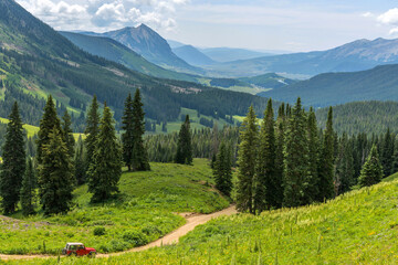 Mount Crested Butte - A red SUV running down a winding mountain road in a green valley towards Mount Crested Butte on a sunny Summer morning. Crested Butte, Colorado, USA.