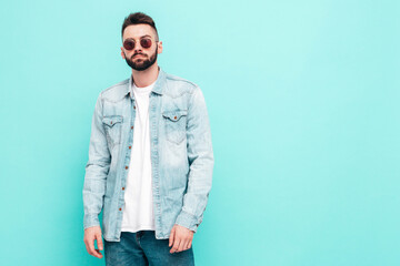 Portrait of handsome confident stylish hipster lambersexual model.Man dressed in jacket and jeans. Fashion male posing near blue wall in studio in sunglasses
