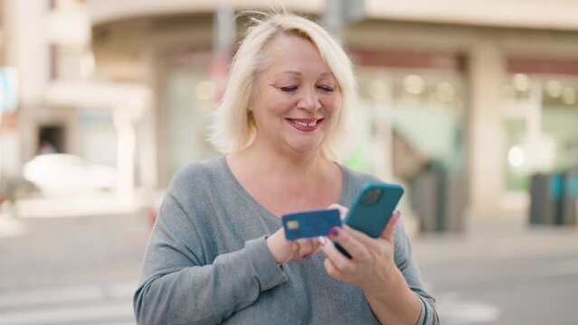 Middle age blonde woman using smartphone and credit card at street