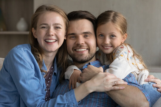Happy family couple of parents and sweet daughter kid hugging, relaxing together at home, looking at camera with toothy smiles. Girl embracing dad from behind. Parenthood concept. Head shot portrait