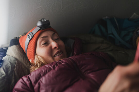 a woman with a headlamp lies in a sleeping bag in a tent and holds a smartphone